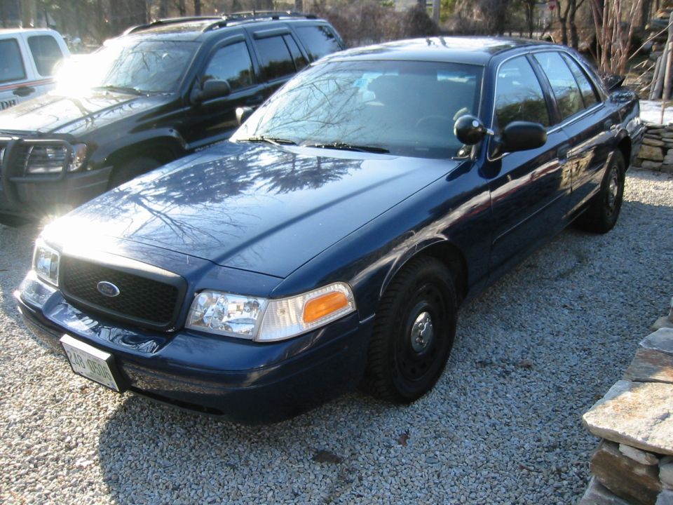 Dark blue 2003 Ford Crown Victoria police car parked on gravel, another SUV behind it