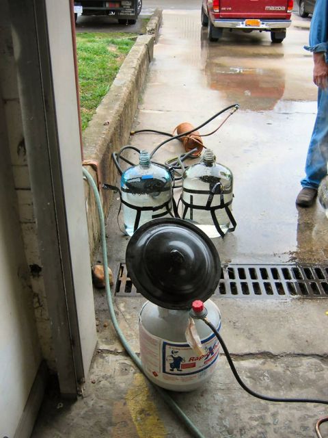 1985 Ford LTD Crown Victoria - Two glass carboys with black straps and hoses are connected to a propane tank with a black lid, set up on a wet concrete surface.