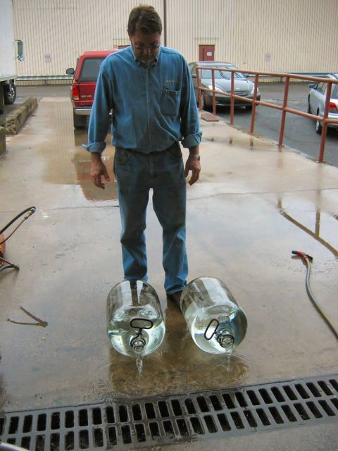 1985 Ford LTD Crown Victoria - A man stands on wet concrete as two large water jugs drain onto the ground, with a red pickup truck in the background.