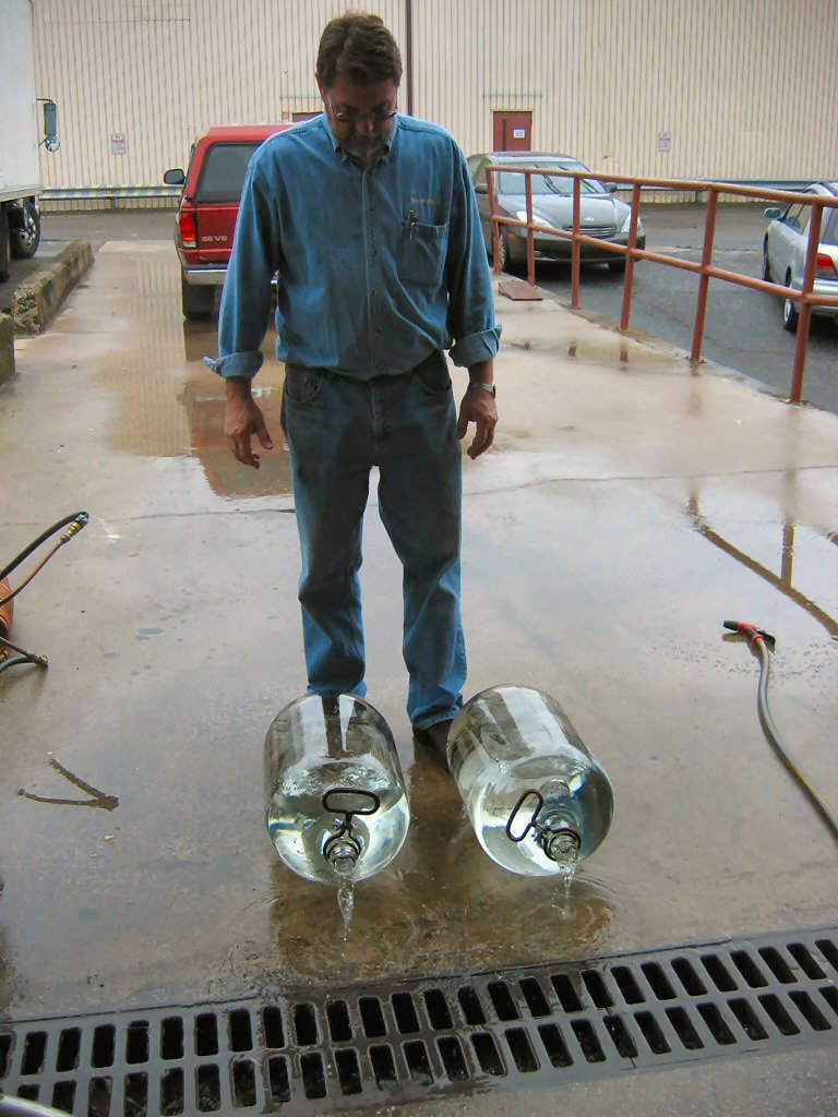 Man standing on wet concrete next to two large glass water jugs draining onto the ground. A red pickup truck is parked behind him