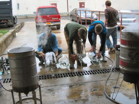 1985 Ford LTD Crown Victoria - Several men are seen draining large glass carboys outdoors, with a red pickup truck and brewing equipment in the background.