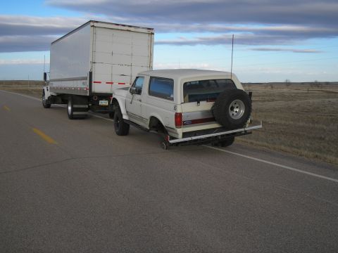 1990 Ford Bronco - A white 1990 Ford Bronco is seen towing a large box truck on a highway under a cloudy sky.
