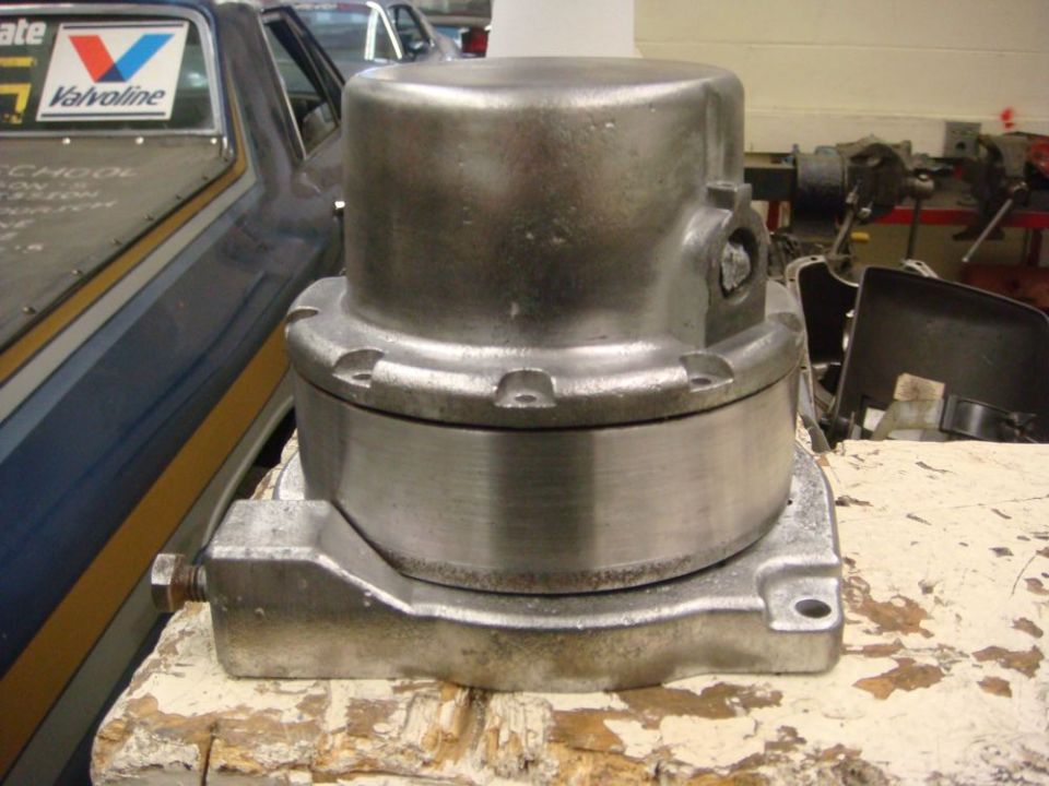 Metal winch mechanism on a workbench, with a blue and yellow striped vehicle in the background