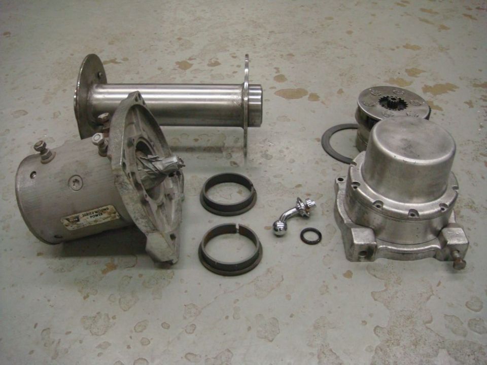 Disassembled winch components laid out on a concrete floor. Includes a drum, motor housing, gears, and other small parts