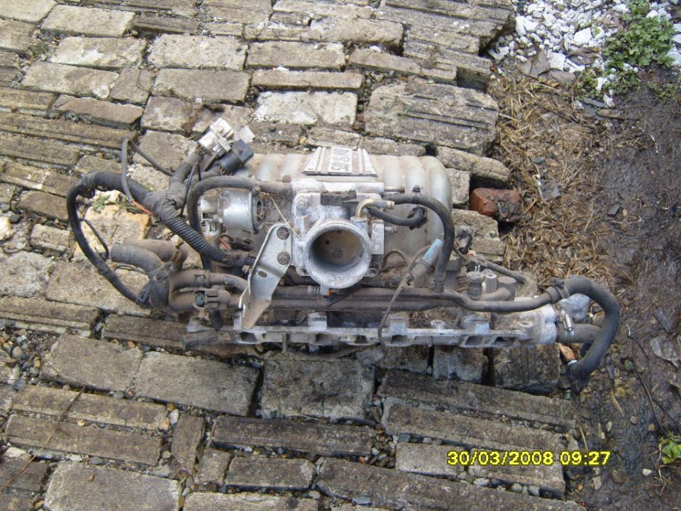Engine intake manifold with throttle body and wiring harness, lying on a brick surface