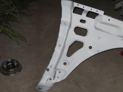 1996 Ford Bronco - A freshly painted white sheet-metal body panel (appears to be an inner fender/apron) resting on a concrete floor, showing multiple stamped…