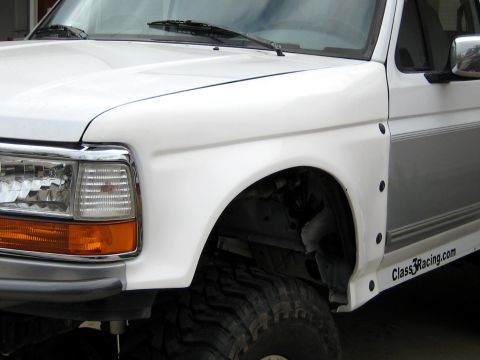1996 Ford Bronco - Front driver-side corner of a white OBS-era Ford truck mid-build, with the fender flare removed to expose the wheel well and lifted suspens…