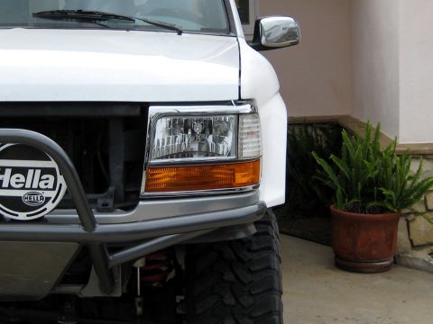 1996 Ford Bronco - Close-up of the driver-side front corner of a lifted white Ford truck/SUV, showing the chrome-trimmed composite headlight with amber turn-s…