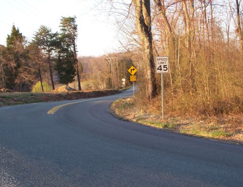 1971 Ford Ranchero - A two-lane rural blacktop road curving through bare winter woods in low golden light, with a double yellow centerline. A "SPEED LIMIT 45" s…