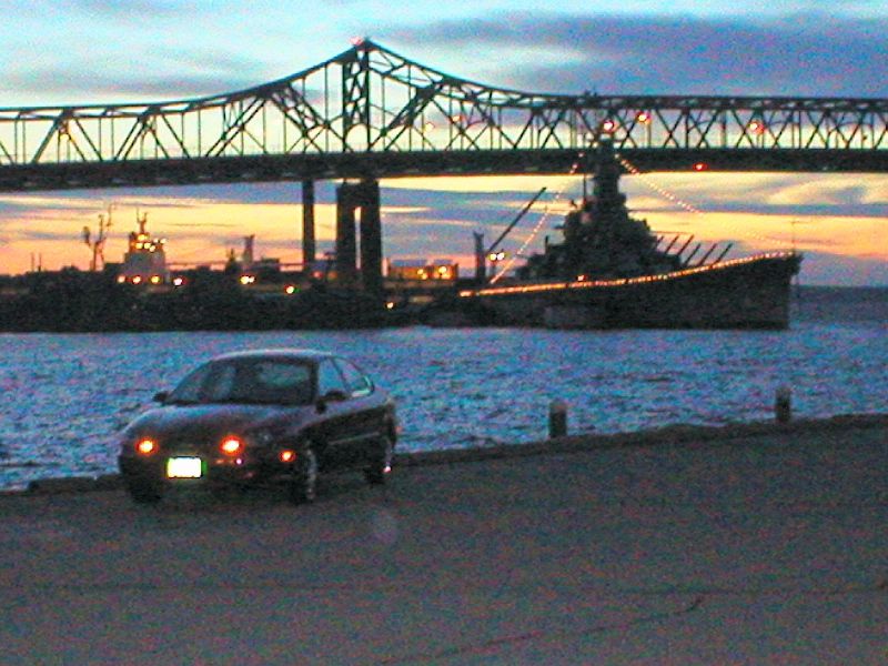 Dark Ford Taurus sedan parked at a waterfront at dusk with a lit battleship and bridge in the background