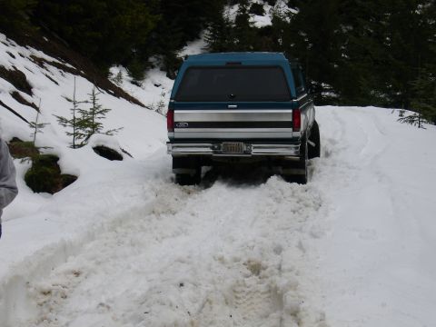 1978 Ford Bronco - A teal Ford Bronco navigates a snowy mountain road, leaving tire tracks in its wake.