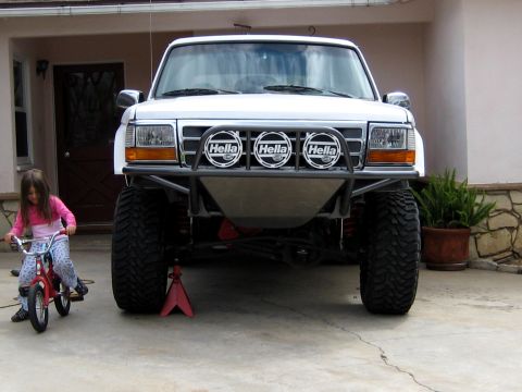 1996 Ford Bronco - Head-on view of a lifted white Ford Bronco on a driveway, fitted with a black tubular prerunner front bumper carrying three round Hella dri…