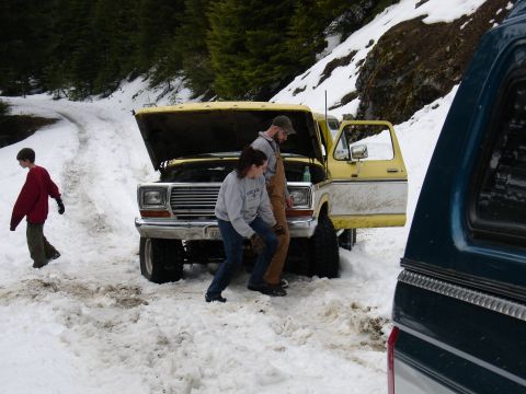 1978 Ford Bronco - Two people check the engine of a yellow Ford F-Series truck stuck in the snow, with a child nearby.