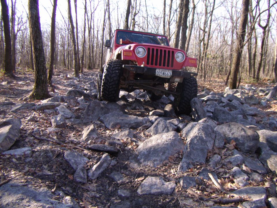 Red 2003 Jeep Wrangler with large tires navigating a rocky trail in a forest
