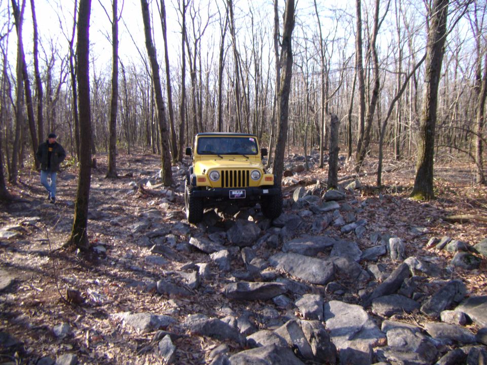 Yellow 2003 Jeep Wrangler on a rocky forest trail, front view. A person walks in the background