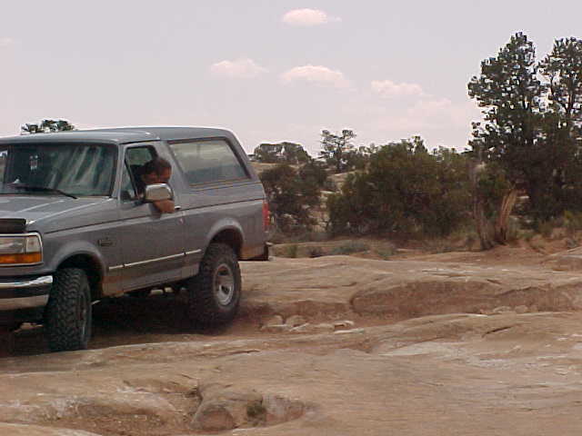 Gray Ford Bronco SUV on slickrock off-road trail, driver's arm out window, desert trees behind