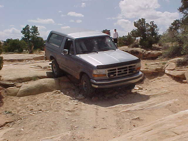 Gray Ford Bronco crawling over rock ledge on slickrock trail, person standing behind