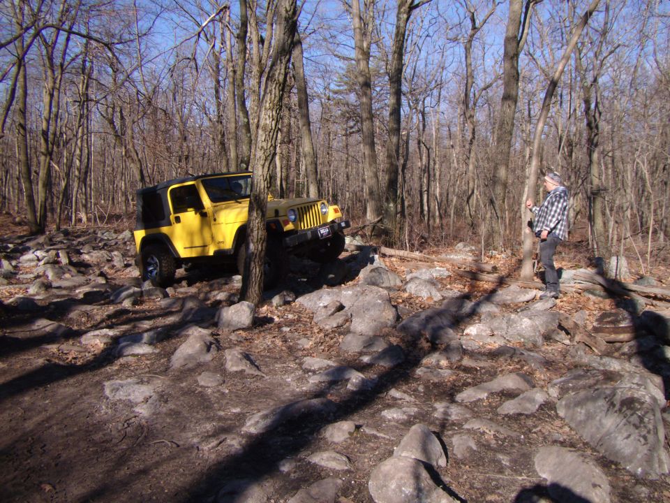 Yellow 2003 Jeep Wrangler with a black soft top navigates a rocky, wooded trail
