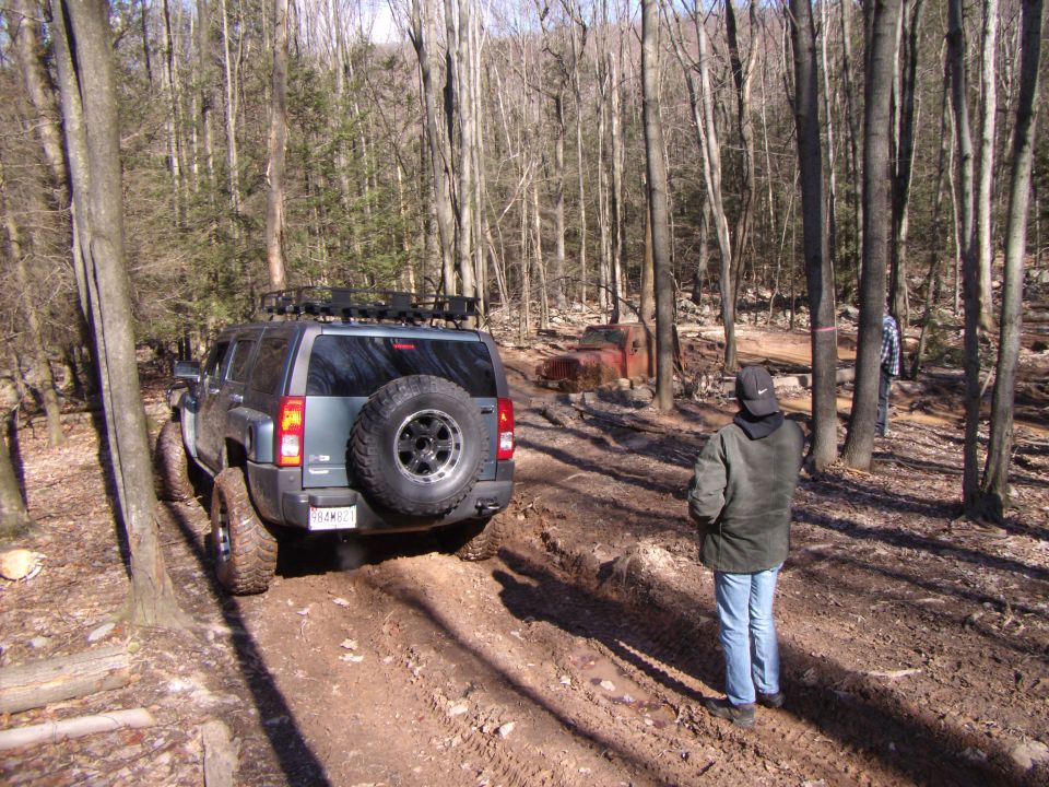 Gray SUV with roof rack and spare tire on rear, on a muddy forest trail. Another rusty vehicle is visible in the background