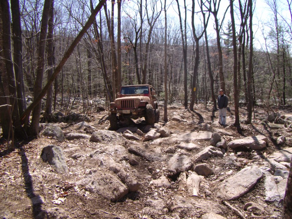 Brown Jeep Wrangler navigating a rocky trail in a wooded area. A person stands to the right