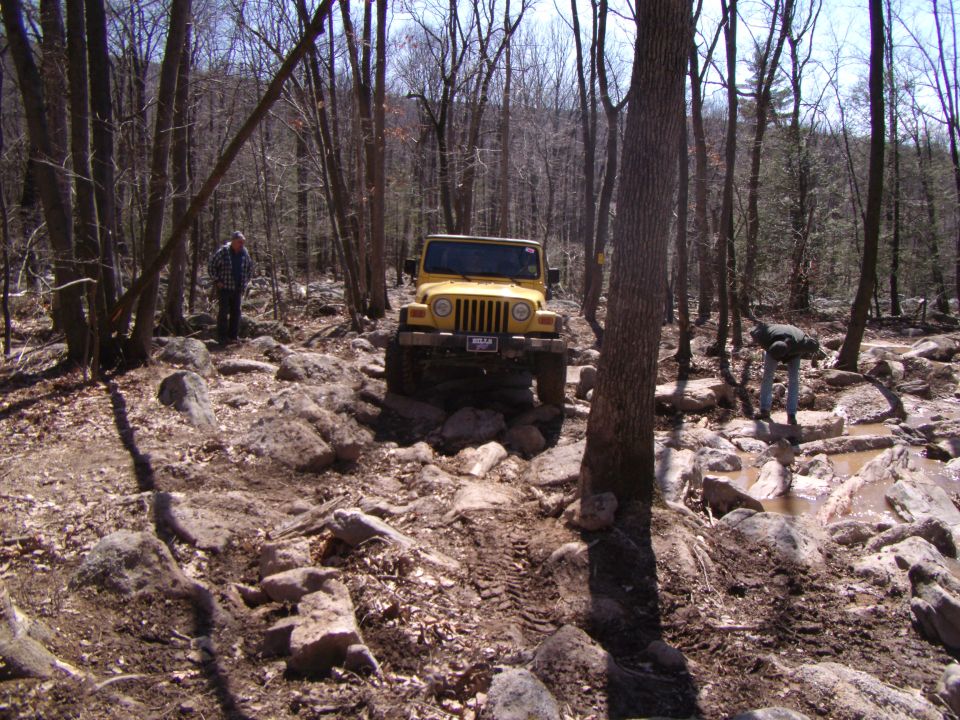 Yellow 2003 Jeep Wrangler on a rocky, wooded trail, front-right quarter view. Muddy tires, some damage visible