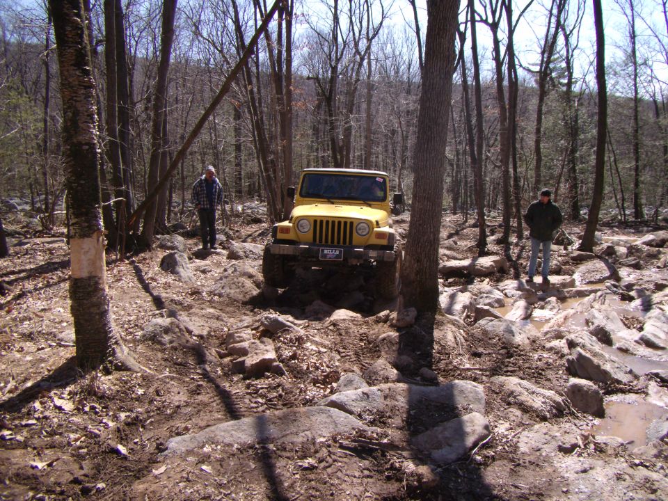 Yellow 2003 Jeep Wrangler navigating a rocky off-road trail with two people standing nearby