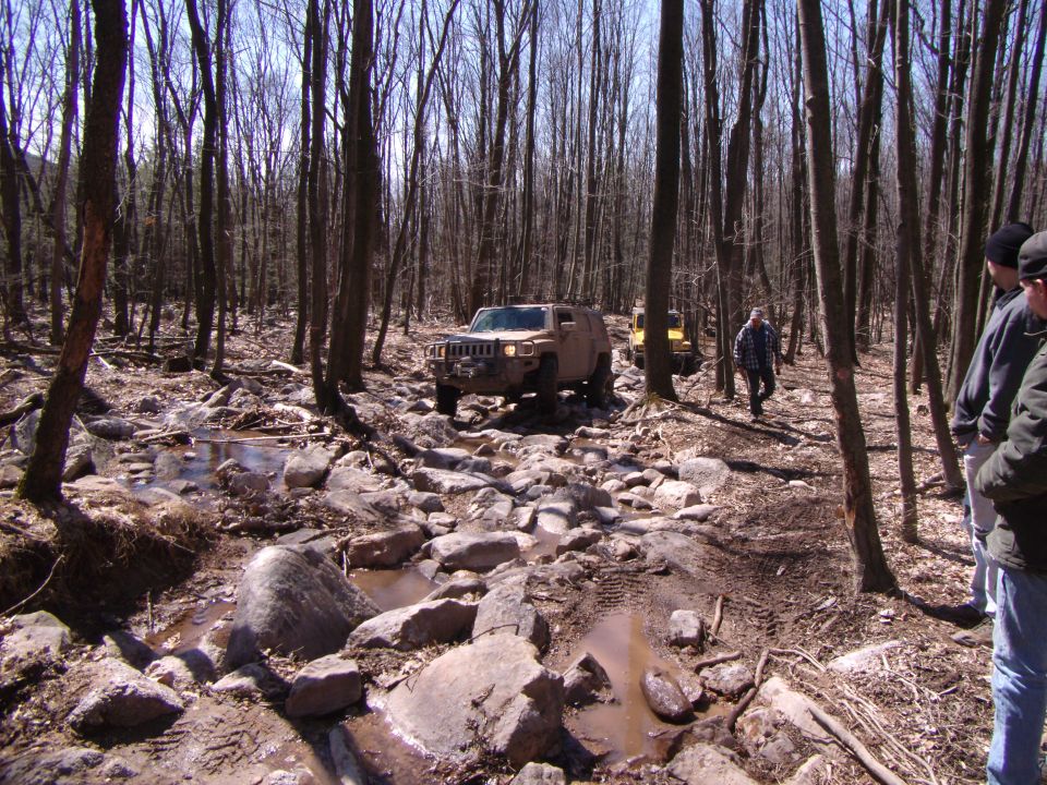 Tan Hummer H3 navigating a rocky, wooded trail with a yellow vehicle behind it