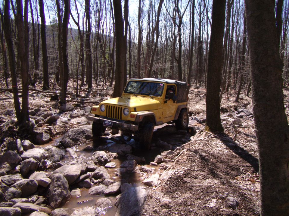 Yellow 2003 Jeep Wrangler driving on a rocky trail in a forest