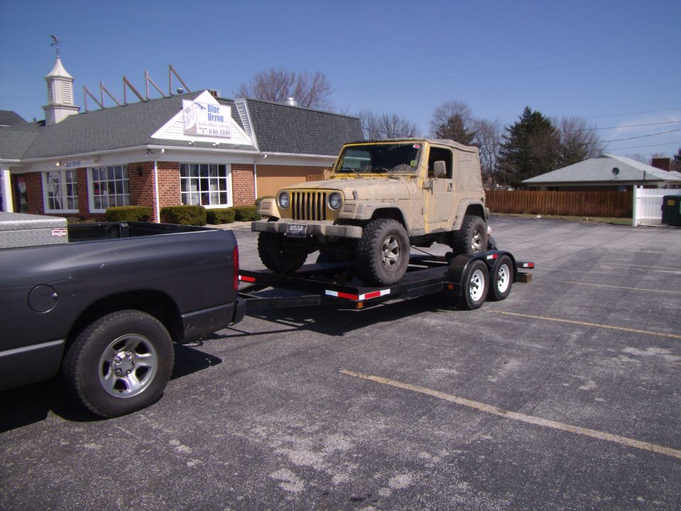 Yellow Jeep Wrangler on a trailer being towed by a dark gray pickup truck in a parking lot