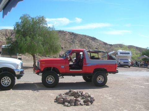 1985 Ford Bronco - A red and grey Ford Bronco is parked on a dirt lot with a white truck and RV in the background.
