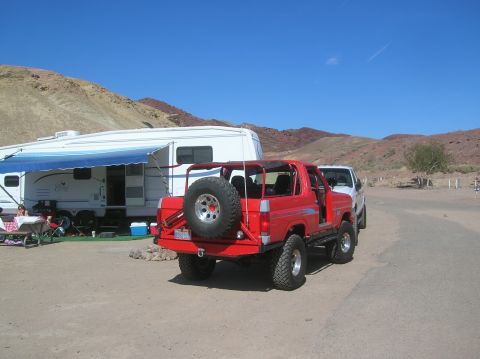 1985 Ford Bronco - A red SUV is parked in a desert campsite next to a large RV and a white truck under a clear blue sky.