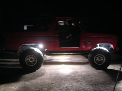 1985 Ford Bronco - A red 1985 Ford Bronco is shown in profile, a lifted suspension and large off-road tires, illuminated by external lights.