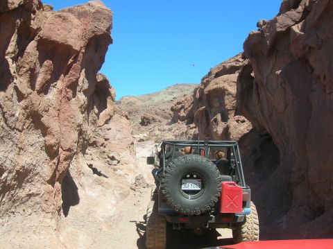 1985 Ford Bronco - A Jeep Wrangler navigates a narrow, rocky canyon trail, with another vehicle visible ahead on the path.
