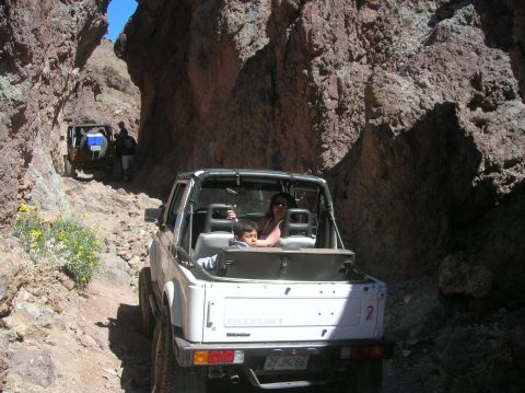 1985 Ford Bronco - Two off-road vehicles navigate a narrow, rocky canyon under a bright blue sky.