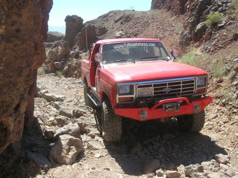 1985 Ford Bronco - A red Ford Bronco navigates a rough, rocky terrain under a clear blue sky.