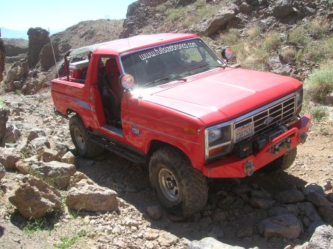 1985 Ford Bronco - This red 1985 Ford Bronco navigates a rocky trail, displaying a website sticker on its windshield.