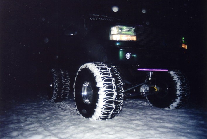 Heavily lifted dark Ford Bronco monster truck on snow at night, low front view