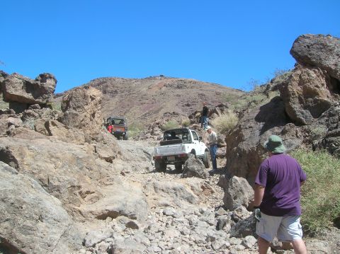 1985 Ford Bronco - Off-road vehicles traverse a challenging, rocky path in a desert landscape with sparse vegetation.