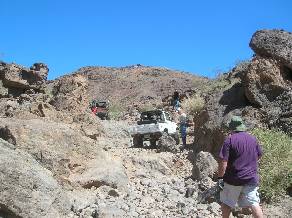 Several off-road vehicles navigate a rocky, desert trail under a clear blue sky. People are visible near the vehicles
