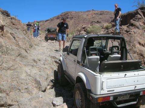 1985 Ford Bronco - A white Suzuki Samurai navigates a challenging rocky terrain with other vehicles and people present.