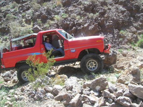 1985 Ford Bronco - A red SUV navigates a rocky, off-road trail with two people inside.