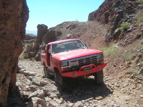 1985 Ford Bronco - A red 1980s Ford Bronco navigates a challenging, rocky off-road path under a clear blue sky.