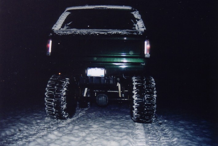 Rear view of a tall-lifted dark green truck on huge mud tires in snow at night