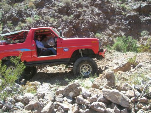 1985 Ford Bronco - Red off-road vehicle with open top navigating rocky terrain, "FSB" visible on fender.