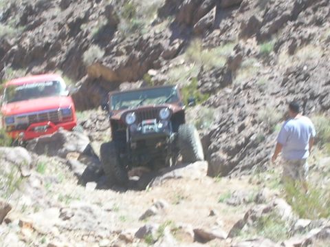 1985 Ford Bronco - Two off-road vehicles navigate a rocky desert landscape, with the red Bronco leading the way.