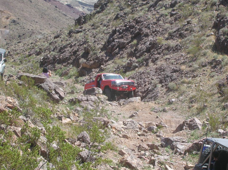 Red pickup truck with off-road modifications navigating a steep, rocky trail. Another vehicle is partially visible in the foreground right