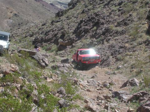 1985 Ford Bronco - Two off-road vehicles navigate a steep, rocky path in a desert canyon.