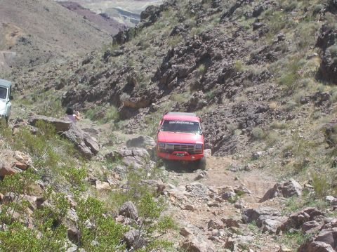 1985 Ford Bronco - A red off-road vehicle navigates a challenging rocky terrain on a sunny day.