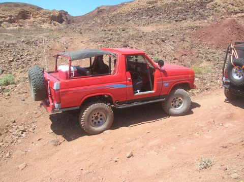 1985 Ford Bronco - Red off-road SUV with a soft top and spare tire on the back is parked on a dirt trail.