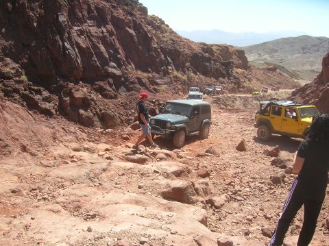 1985 Ford Bronco - Multiple Jeeps navigate a challenging off-road trail through a rocky canyon under a clear blue sky.